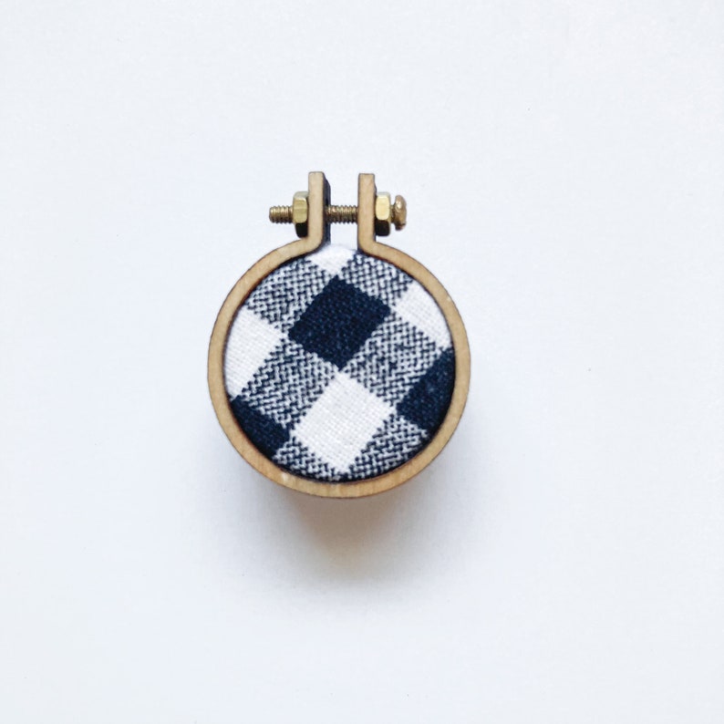 May include: A small wooden embroidery hoop with a black and white checked fabric inside. The hoop is approximately 5 cm in diameter.