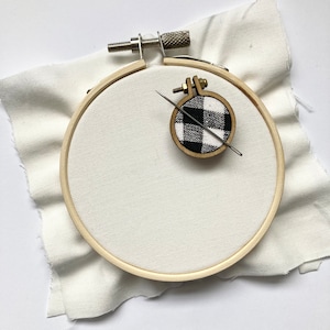 May include: A white embroidery hoop with a black and white checked fabric circle inside. A needle is threaded through the fabric.
