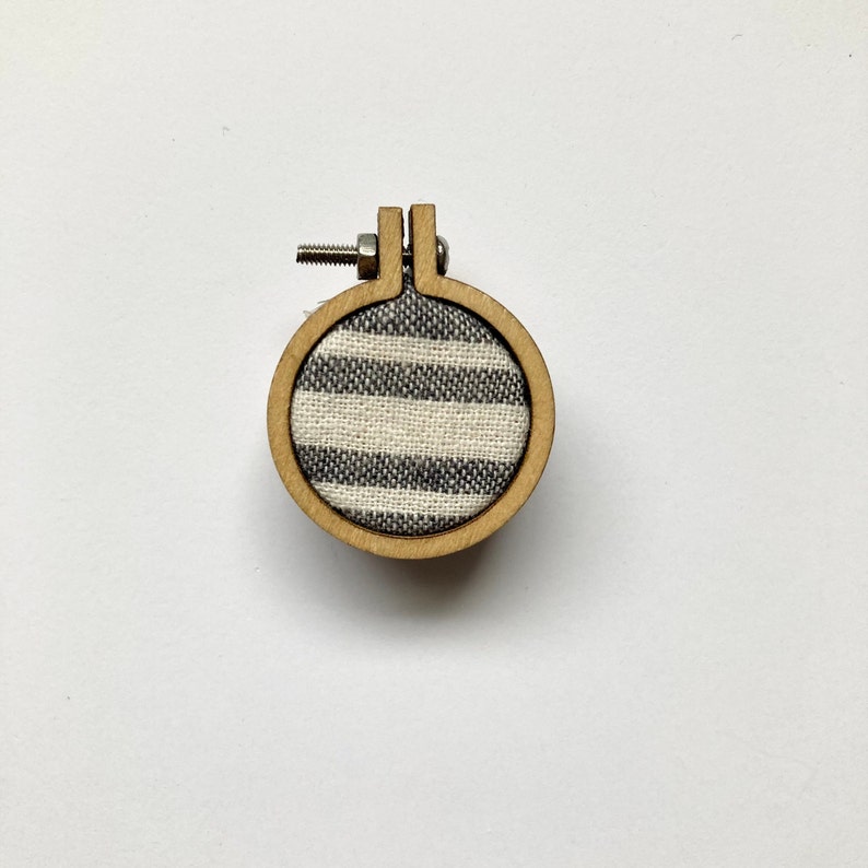 May include: A small, round wooden embroidery hoop with a white and grey striped fabric inside. The hoop is approximately 5 cm in diameter.