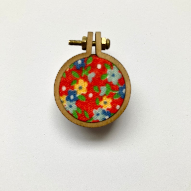 May include: A small wooden embroidery hoop with a red fabric background and a floral pattern in blue, yellow, and green.