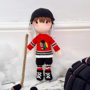 Op de afbeelding: Een handgemaakte gehaakte pop verkleed als een hockeyspeler. De pop draagt een rood shirt met een teamlogo, een zwarte broek en een zwarte helm. Een hockeystick en puck zijn inbegrepen. De pop is ongeveer 20 cm hoog.