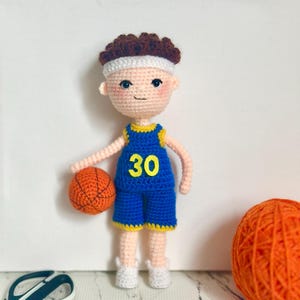 Puede incluir: Muñeco de ganchillo de jugador de baloncesto con uniforme azul y amarillo, número 30. El muñeco tiene pelo castaño, una cinta blanca y sostiene una pelota de baloncesto naranja. Zapatos blancos.