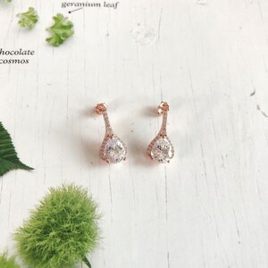 Op de afbeelding: Een paar ros&eacute;gouden druppelvormige oorbellen met een heldere, sprankelende middensteen en een halo van kleinere stenen. De oorbellen hebben een delicaat, langwerpig ontwerp. Ze liggen op een witte houten ondergrond. Een elegant sieraad.