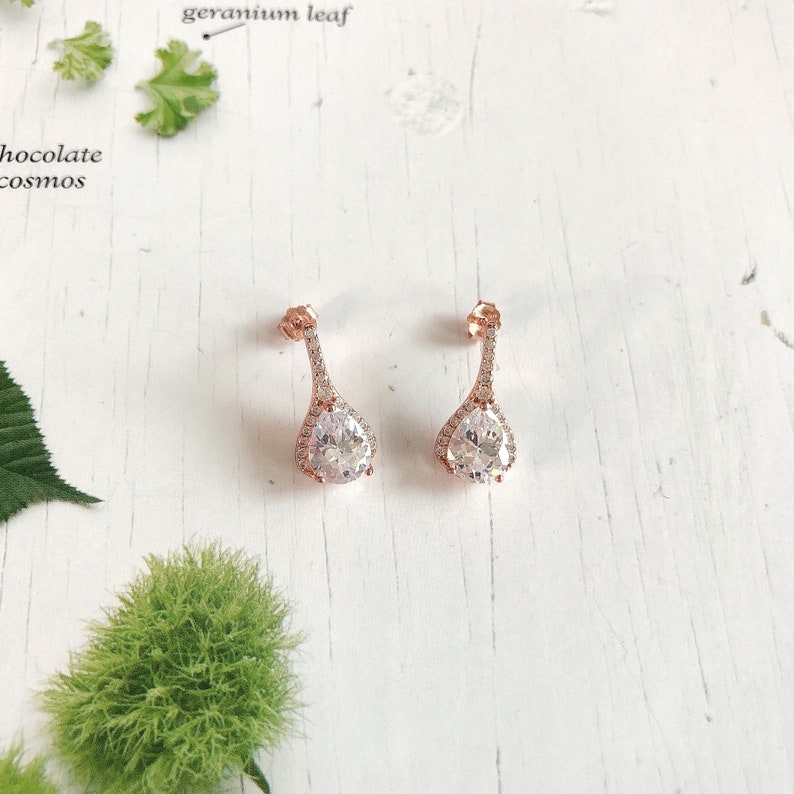 Op de afbeelding: Een paar ros&eacute;gouden druppelvormige oorbellen met een heldere, sprankelende middensteen en een halo van kleinere stenen. De oorbellen hebben een delicaat, langwerpig ontwerp. Ze liggen op een witte houten ondergrond. Een elegant sieraad.