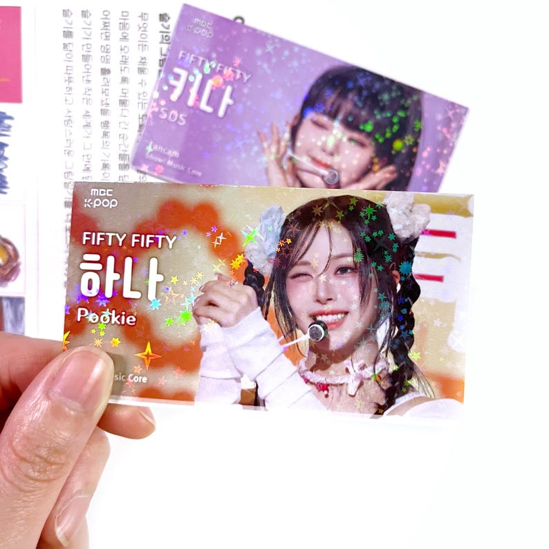 May include: Two holographic stickers featuring a K-pop singer. The bottom sticker has the text "FIFTY FIFTY" in English and Korean, with the name "하나" and "Pookie". The top sticker has the text "FIFTY FIFTY" and Korean text.