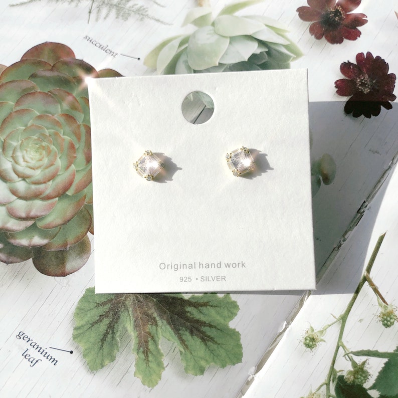 May include: A pair of square-shaped stud earrings with a clear, faceted center stone and gold-colored metal accents. The earrings are displayed on a white card with the text "Original hand work 925 SILVER". The background includes floral and plant elements.