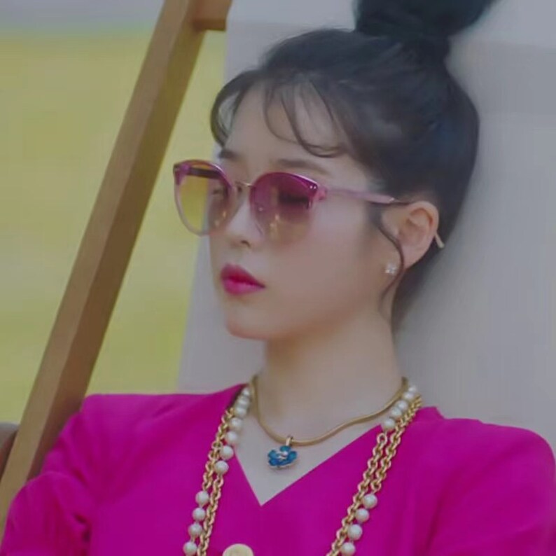 May include: Pink cat-eye sunglasses with gradient lenses. A woman is wearing a gold necklace with a blue pendant and a pearl necklace. She is wearing a bright pink top. Her hair is up in a bun.
