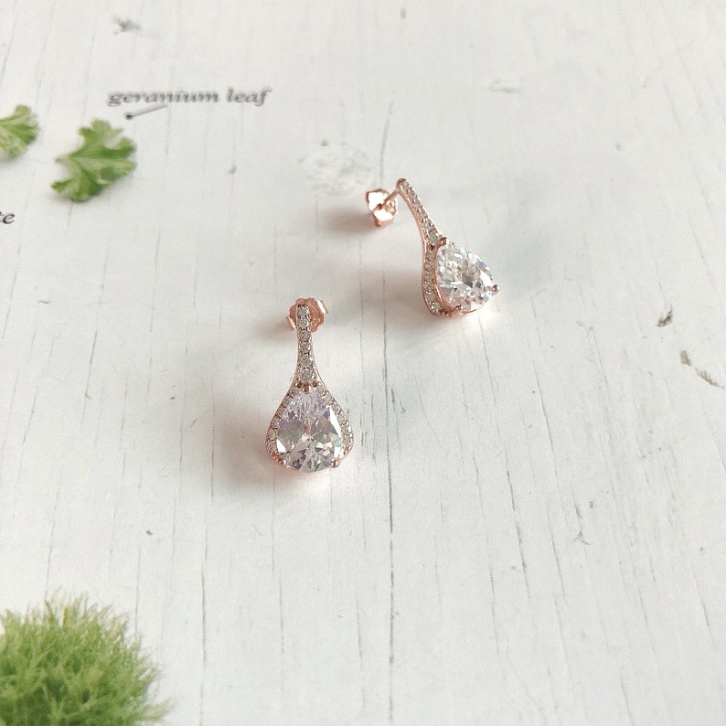 Op de afbeelding: Een paar ros&eacute;gouden oorhangers met een peervormige, heldere kristallen steen. De oorbellen hebben een halo van kleinere kristallen rond de hoofdsteen en langs de pin. Gepresenteerd op een wit houten oppervlak.