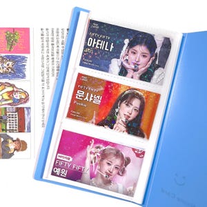 May include: A blue card holder with three photo card slots, each containing a different image of a female K-pop singer. The cards have Korean text and the group name "FIFTY FIFTY". The holder is open, revealing the cards.