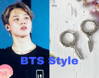 May include: A pair of silver hoop earrings with a feather charm hanging from each hoop. The earrings are on a white wooden surface with pink flowers.