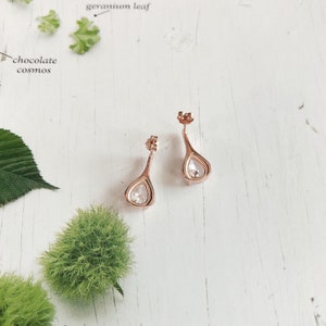 Op de afbeelding: Een paar ros&eacute;gouden druppelvormige oorbellen met heldere, sprankelende stenen. De oorbellen worden getoond op een witte, getextureerde ondergrond met botanische elementen op de achtergrond. De oorbellen hebben een steker.