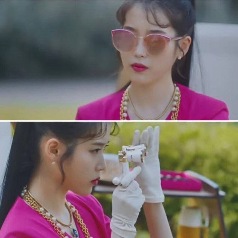 May include: Pink cat-eye sunglasses with a gold frame. The image also shows a gold necklace with pearls and a white and gold object being held by a person wearing white gloves.