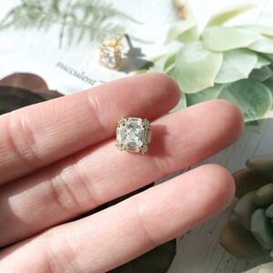May include: Close-up of a gold-toned square stud earring with a large, clear, square-cut stone at the center. The earring is surrounded by smaller, clear stones. A second earring is visible in the background.
