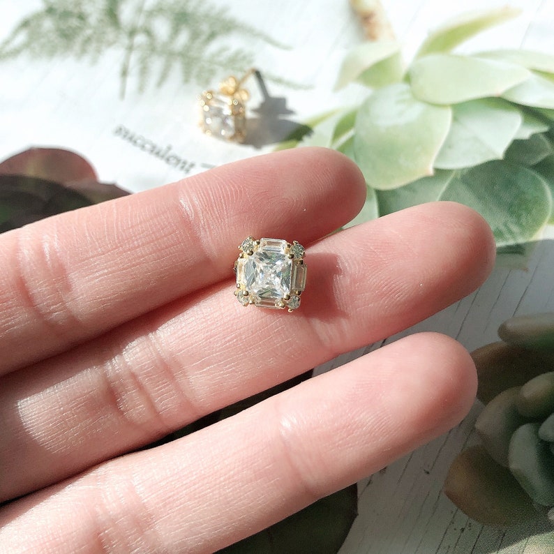 May include: Close-up of a gold-toned square stud earring with a large, clear, square-cut stone at the center. The earring is surrounded by smaller, clear stones. A second earring is visible in the background.