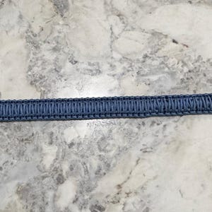 May include: A long, narrow, blue paracord strap featuring a tightly woven, textured pattern. The strap is finished with dark metal hardware at both ends, including small, integrated swivel lobster claw clasps for secure attachment.