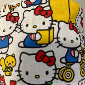 May include: White fabric with a repeating pattern of Hello Kitty characters in various poses. The characters are in shades of red, blue, yellow, and black on a white background.