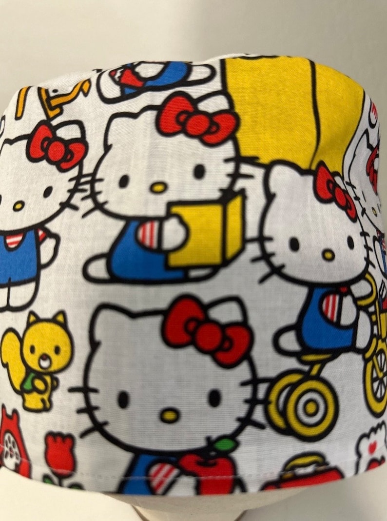 May include: White fabric with a repeating pattern of Hello Kitty characters in various poses. The characters are in shades of red, blue, yellow, and black on a white background.