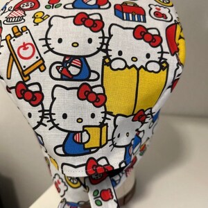 May include: White fabric scrub hat with a colorful pattern of Hello Kitty characters, books, and school supplies. The hat has a tie at the back.