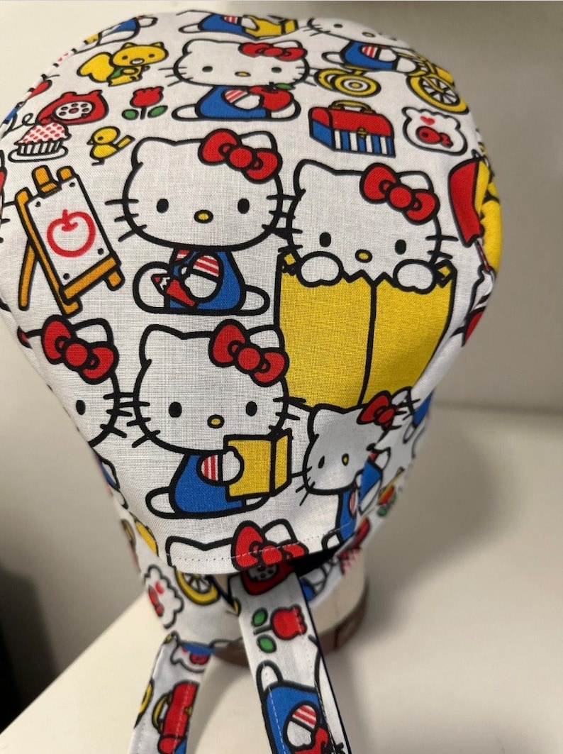 May include: White fabric scrub hat with a colorful pattern of Hello Kitty characters, books, and school supplies. The hat has a tie at the back.