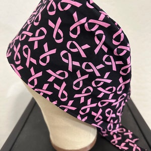 May include: Black scrub hat with a pink ribbon pattern. The hat has a long tie that can be tied at the back of the neck.