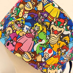 May include: A blue and yellow surgical scrub hat with a colorful print of various characters from the Super Mario video game franchise.