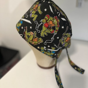 May include: Black scrub hat with a repeating pattern of Teenage Mutant Ninja Turtles characters and white stars. The hat has two long ties.
