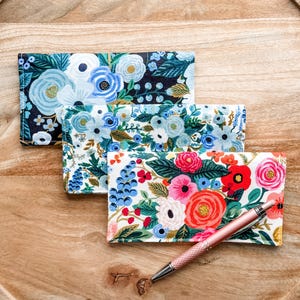 May include: Three floral print card holders in blue, white, and red hues are displayed on a wooden tray. A rose gold pen rests on the tray, next to the card holders. The card holders feature various floral patterns.