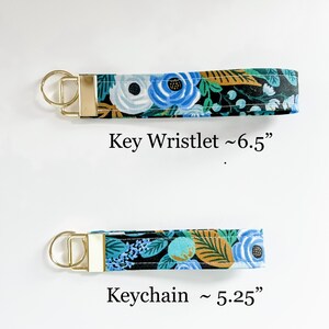 May include: Two fabric keychains with gold metal rings. The top keychain is a wristlet style and measures approximately 6.5 inches long. The bottom keychain is a shorter style and measures approximately 5.25 inches long. Both keychains feature a floral pattern with blue, white, green, and gold accents on a black background.