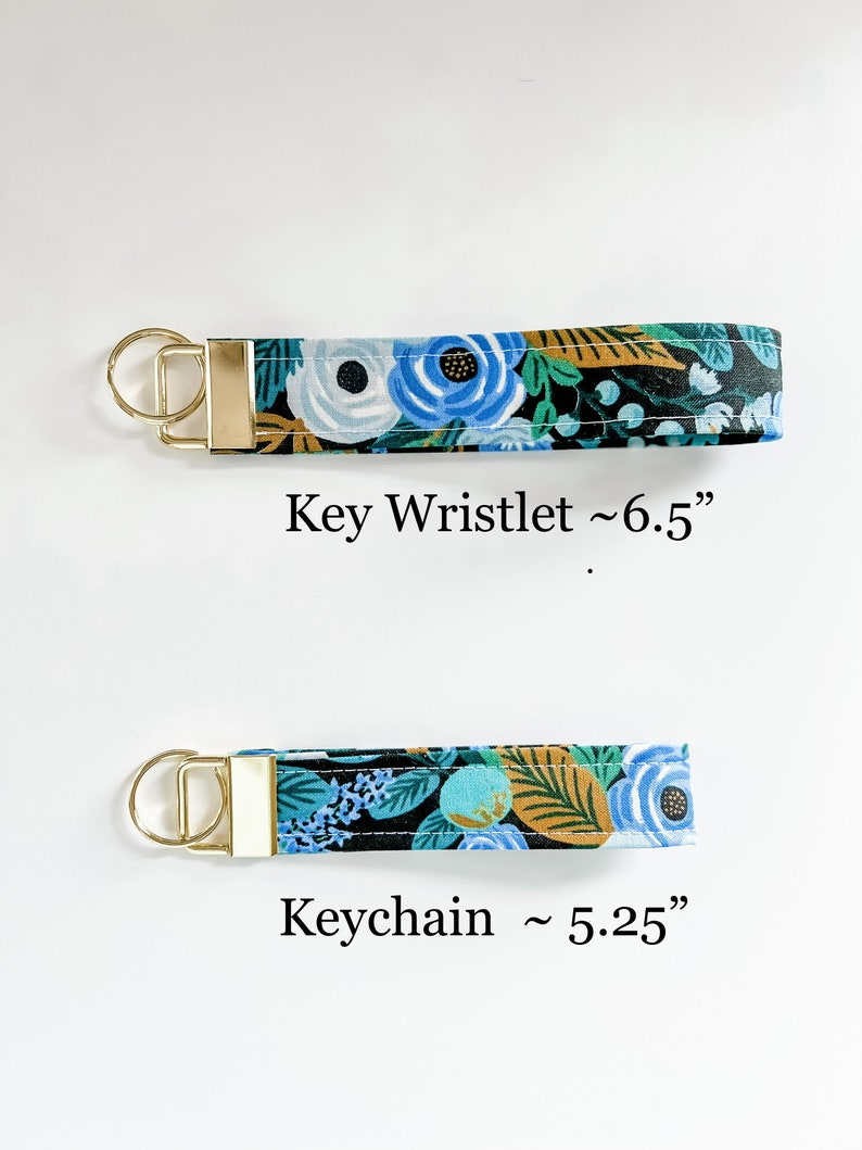 May include: Two fabric keychains with gold metal rings. The top keychain is a wristlet style and measures approximately 6.5 inches long. The bottom keychain is a shorter style and measures approximately 5.25 inches long. Both keychains feature a floral pattern with blue, white, green, and gold accents on a black background.
