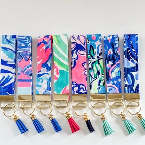 May include: A collection of eight wrist keychains with colorful patterned fabric straps. Each keychain features a gold-colored metal ring and a matching tassel in shades of blue, pink, and green. The fabric patterns include floral and abstract designs.