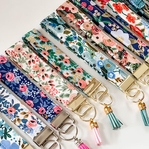 May include: A collection of floral patterned key fobs in various colors. Each fabric wristlet features a gold or silver-toned metal clasp and a matching tassel. The designs include pink, blue, and red flowers on navy, white, and green backgrounds.