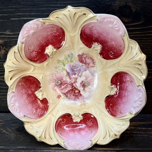May include: A vintage porcelain bowl with a scalloped edge and a floral design. The bowl is decorated with pink, red, and gold accents. The floral design features pink and purple flowers with green leaves.