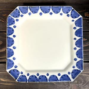 May include: Square white ceramic plate with a blue scalloped border design. The plate has an octagonal shape with a smooth, glossy finish. The blue border features a repeating pattern of stylized cloud-like shapes.
