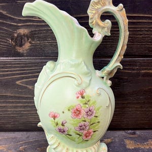 May include: A light green ceramic pitcher with a gold-accented handle and floral design. The pitcher has a scalloped base and a delicate floral pattern.