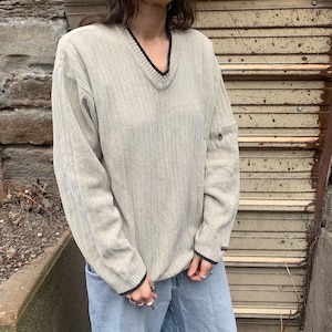 May include: A light gray ribbed knit sweater with a black v-neck and black trim on the sleeves. The sweater has a zippered pocket on the left sleeve.