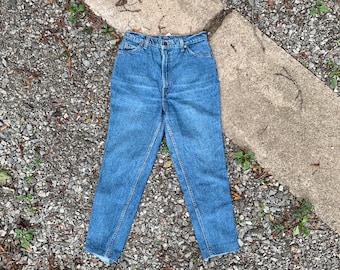 May include: A pair of blue denim jeans with a high waist and a straight leg. The jeans have a faded look and are laid flat on a gravel surface.