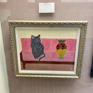 May include: Framed painting titled "Two Muses" featuring a gray cat seated on a pink sofa next to a yellow vase with red flowers. The painting has a gold ornate frame and is displayed on a pink wall.