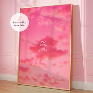 Op de afbeelding: Een ingelijste print met een roze lucht met wolken. De tekst "GOOD THINGS ARE COMING" staat in het midden van de print. De lijst is goudkleurig en de print leunt tegen een roze muur. Een witte cirkel met de tekst "Personalise Your Print" staat linksboven.