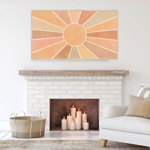 Puede incluir: Una obra de arte abstracta con forma de sol radiante, en color beige y naranja, cuelga sobre una chimenea de ladrillo blanco con velas encendidas en su interior. La obra de arte est&aacute; enmarcada en un marco de madera clara.