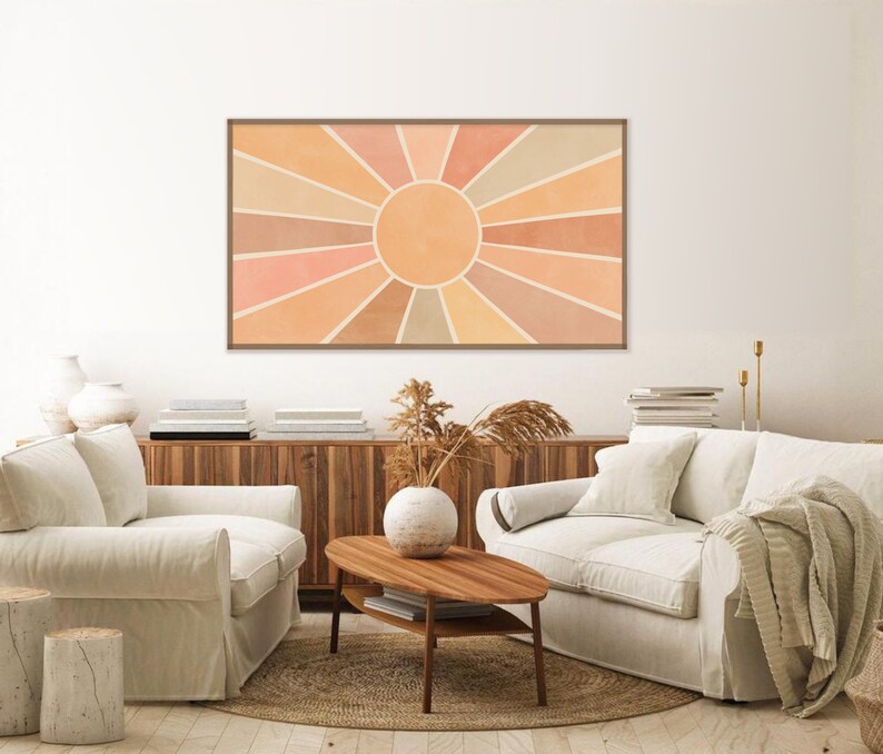 Puede incluir: Un dise&ntilde;o de interior de sala de estar con dos sof&aacute;s blancos, una mesa de caf&eacute; de madera y una impresi&oacute;n enmarcada de un dise&ntilde;o de sol en forma de rayos en tonos naranja, melocot&oacute;n y beige. La impresi&oacute;n est&aacute; colgada en una pared blanca sobre un gabinete de madera.