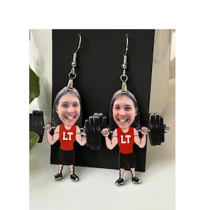 May include: A pair of dangle earrings featuring a cartoon illustration of a person with a smiling face wearing a red tank top with the letters LT in white, black shorts, and black shoes, while lifting a black barbell with weights on each side.
