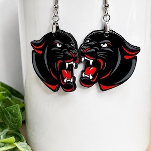 May include: A pair of dangle earrings featuring black panther heads with snarling expressions, white fangs, and vibrant red accents inside the open mouths and along the ears. The earrings hang from silver-toned metal hooks and jump rings.