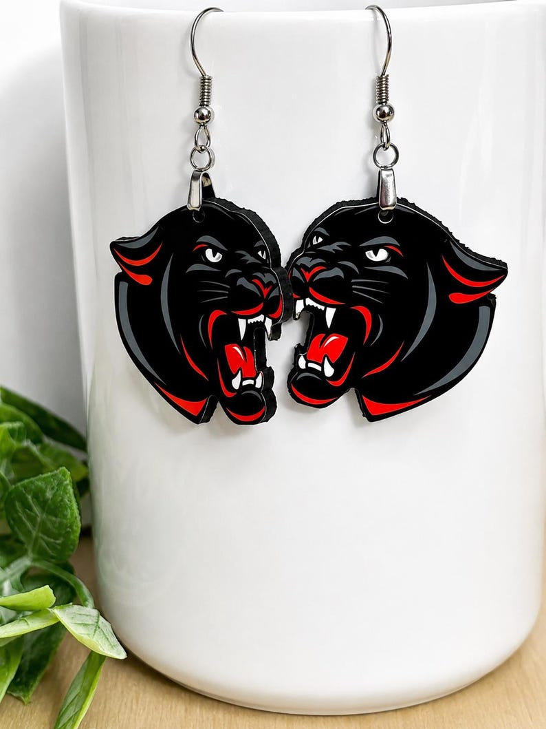 May include: A pair of dangle earrings featuring black panther heads with snarling expressions, white fangs, and vibrant red accents inside the open mouths and along the ears. The earrings hang from silver-toned metal hooks and jump rings.