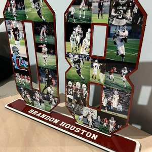 Puede incluir: Un n&uacute;mero 8 de madera rojo y blanco con un collage de fotos de un jugador de f&uacute;tbol americano con un uniforme granate y blanco. La base del n&uacute;mero es roja con letras blancas que dicen "BRANDON HOUSTON".