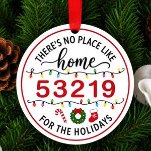 以下が含まれることがあります： 赤い縁取りと赤いリボンが付いた白い円形のクリスマスオーナメント。黒と赤で「THERE'S NO PLACE LIKE home 53219 FOR THE HOLIDAYS」という文字が書かれており、カラフルなイルミネーションライト、キャンディケイン、リース、靴下のイラストで装飾されています。