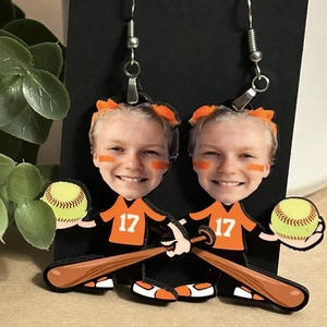 May include: A pair of dangle earrings featuring a custom cutout of a smiling person wearing an orange jersey with the number 17, holding a yellow softball and a brown baseball bat. The earrings have silver-toned metal hooks for attachment.