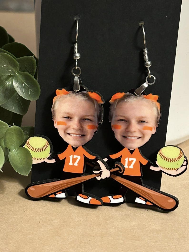 May include: A pair of dangle earrings featuring a custom cutout of a smiling person wearing an orange jersey with the number 17, holding a yellow softball and a brown baseball bat. The earrings have silver-toned metal hooks for attachment.