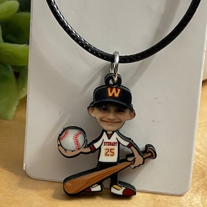 May include: A necklace featuring a custom pendant shaped like a cartoon baseball player wearing a black cap with a yellow W, a white jersey with the name STEWART and number 25, holding a baseball and a wooden bat, suspended on a black cord.