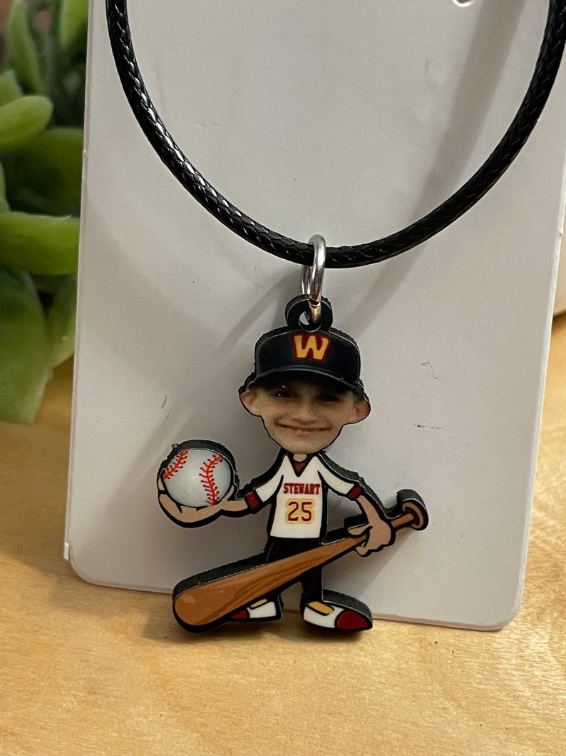 May include: A necklace featuring a custom pendant shaped like a cartoon baseball player wearing a black cap with a yellow W, a white jersey with the name STEWART and number 25, holding a baseball and a wooden bat, suspended on a black cord.