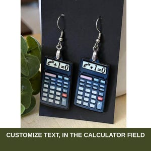 May include: A pair of dangle earrings featuring miniature dark blue calculator charms. Each calculator displays the mathematical equation e^ix+1=0 on its screen. The earrings are attached to silver-toned metal hooks and are presented on a black card.